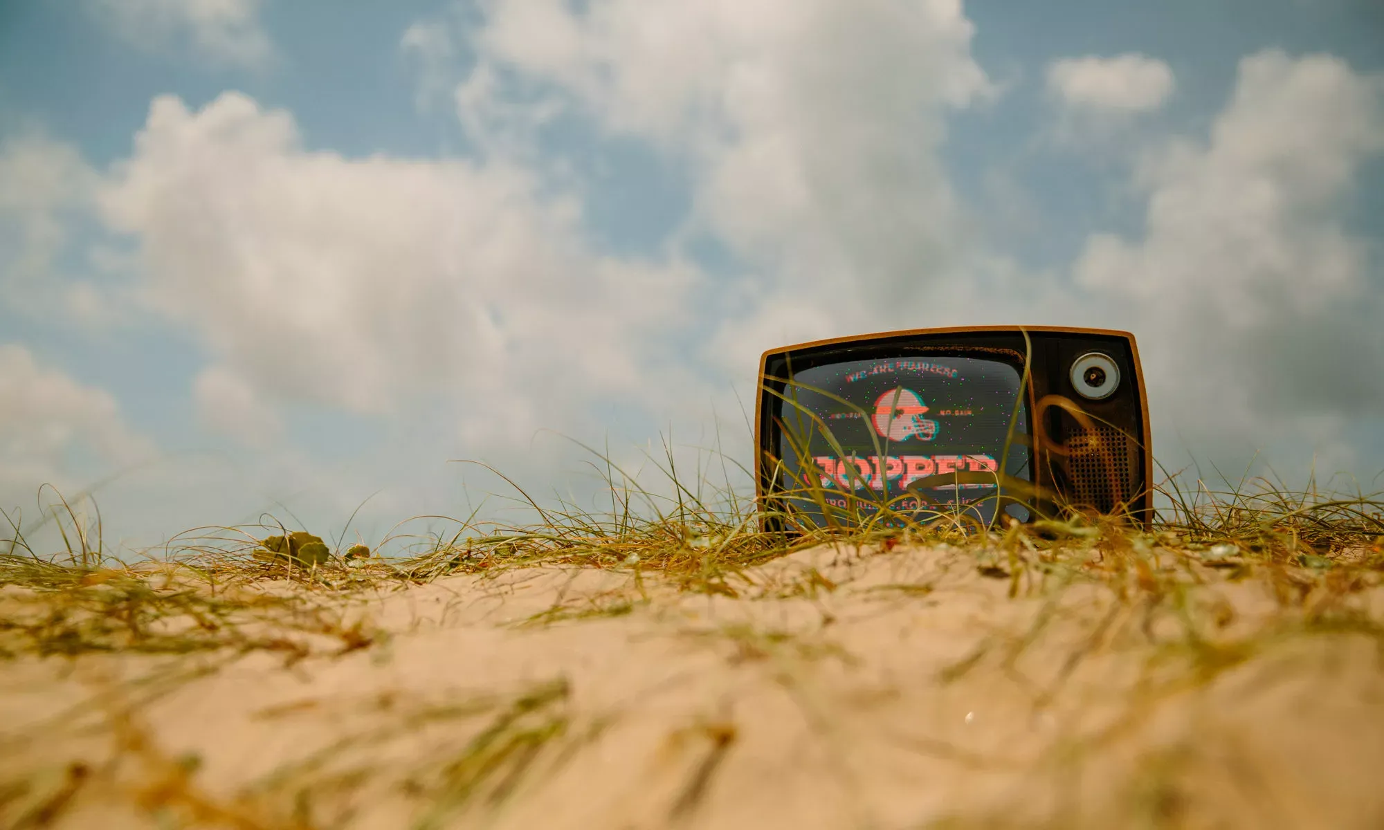 Televisor retro de los setenta semienterrado en la hierba de una duna, con el logo de Jopper en pantalla